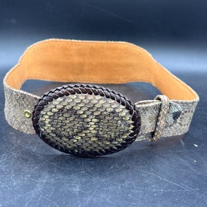 May include: A wide belt featuring a textured, snakeskin-patterned surface in shades of brown and tan. The belt includes a large, oval-shaped buckle with a dark, braided border and a central snakeskin-like inlay. The interior lining is tan.