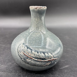 May include: A small, round, blue-grey ceramic vase with a crackle glaze. The vase features a raised feather design on the front and a textured, brown rim. The vase is approximately 5 inches tall.