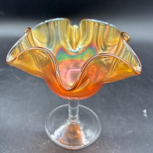 May include: An iridescent orange glass bowl with a ruffled rim, set on a clear glass stem. The bowl's color shifts with light, displaying hues of gold, red, and green. The stem is clear, and the bowl has a decorative, vintage aesthetic.
