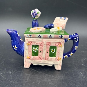 May include: A whimsical ceramic teapot shaped like a miniature kitchen cabinet. The teapot features a blue spout and handle adorned with white flowers. The cabinet is painted pink and white with green accents, and topped with a green surface.