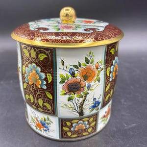 May include: A decorative tin with a lid, featuring a floral design in shades of orange, blue, and white. The lid and base have a dark brown background with gold accents. The tin has a gold knob on top.