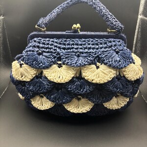 May include: A structured handbag crafted from woven raffia-like material in alternating rows of navy blue and cream fan-shaped patterns. The bag features a single top handle, a dark blue frame, and a gold-toned metal kiss-lock clasp closure.
