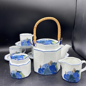 May include: A white ceramic tea set with blue and green tree designs. The set includes a teapot with a woven handle, a creamer, a sugar bowl, cups, and saucers. The teapot is approximately 8 inches tall.