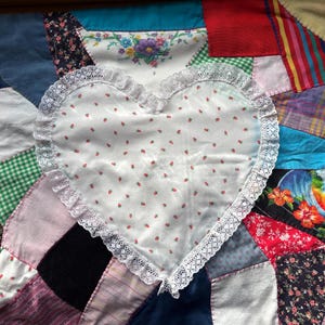 May include: A white heart-shaped pillow with a lace trim is placed on a patchwork quilt made of various fabrics and colors. The quilt is made of squares and rectangles of different sizes and patterns.