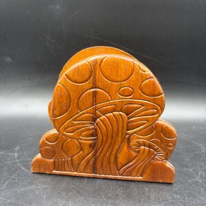 May include: A brown wooden napkin holder featuring a carved relief design of two mushrooms with rounded caps and visible gills on the stems. The wood has a smooth, polished finish with a natural grain pattern visible across the surface.