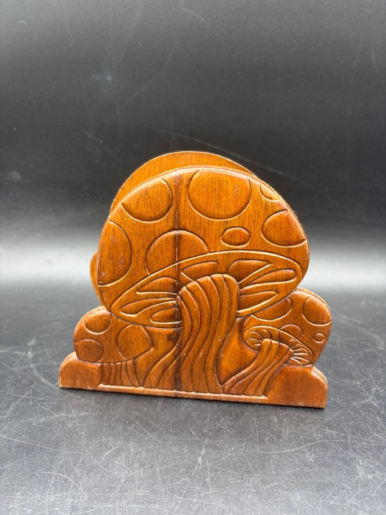May include: A brown wooden napkin holder featuring a carved relief design of two mushrooms with rounded caps and visible gills on the stems. The wood has a smooth, polished finish with a natural grain pattern visible across the surface.