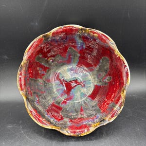 May include: A ceramic bowl with a scalloped rim featuring a glossy glaze in shades of deep red, charcoal gray, and hints of blue. The interior surface displays a marbled, swirling pattern where the colors blend together in a fluid, organic design.