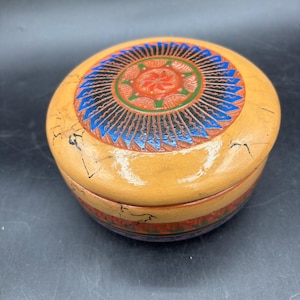 May include: A round wooden lidded box with a glossy tan finish and black crackle detailing. The lid features a central orange circular motif with a star pattern, surrounded by a ring of blue and black radiating lines and green accents.