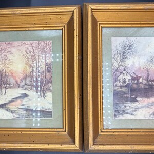 May include: Two framed watercolor paintings depicting winter scenes. The left painting shows a snowy path leading to a small village with a river in the foreground. The right painting shows a bridge over a river with a house and trees in the background.