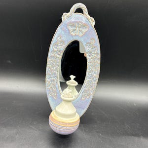 May include: A ceramic wall mirror with a floral design and a butterfly. The mirror is oval shaped and has a small ceramic oil lamp hanging below it.