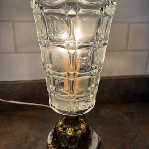 May include: A clear glass lamp with a geometric pattern and a gold metal base. The lamp is on a white marble base.