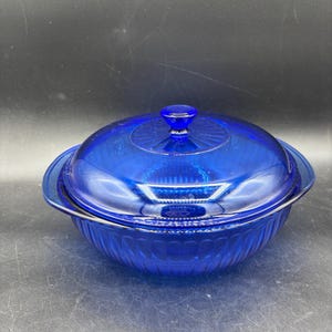 May include: A round, cobalt blue glass casserole dish with a matching lid. The bowl and lid feature a vertical ribbed pattern along the sides, and the lid includes a central knob handle with a sunburst design radiating from the center.