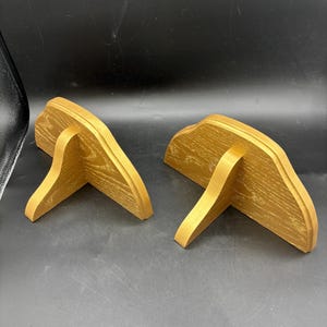 May include: Two wooden wall shelves with a light brown finish. Each shelf has a curved top and a single support leg. The shelves are designed to be mounted on a wall and provide a decorative storage solution.