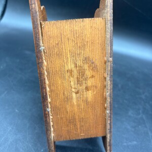 May include: A small, rectangular wooden box with a medium brown finish. The item features two vertical side panels with curved top edges and a central compartment. The wood grain is visible throughout, showing natural variations in color and texture.
