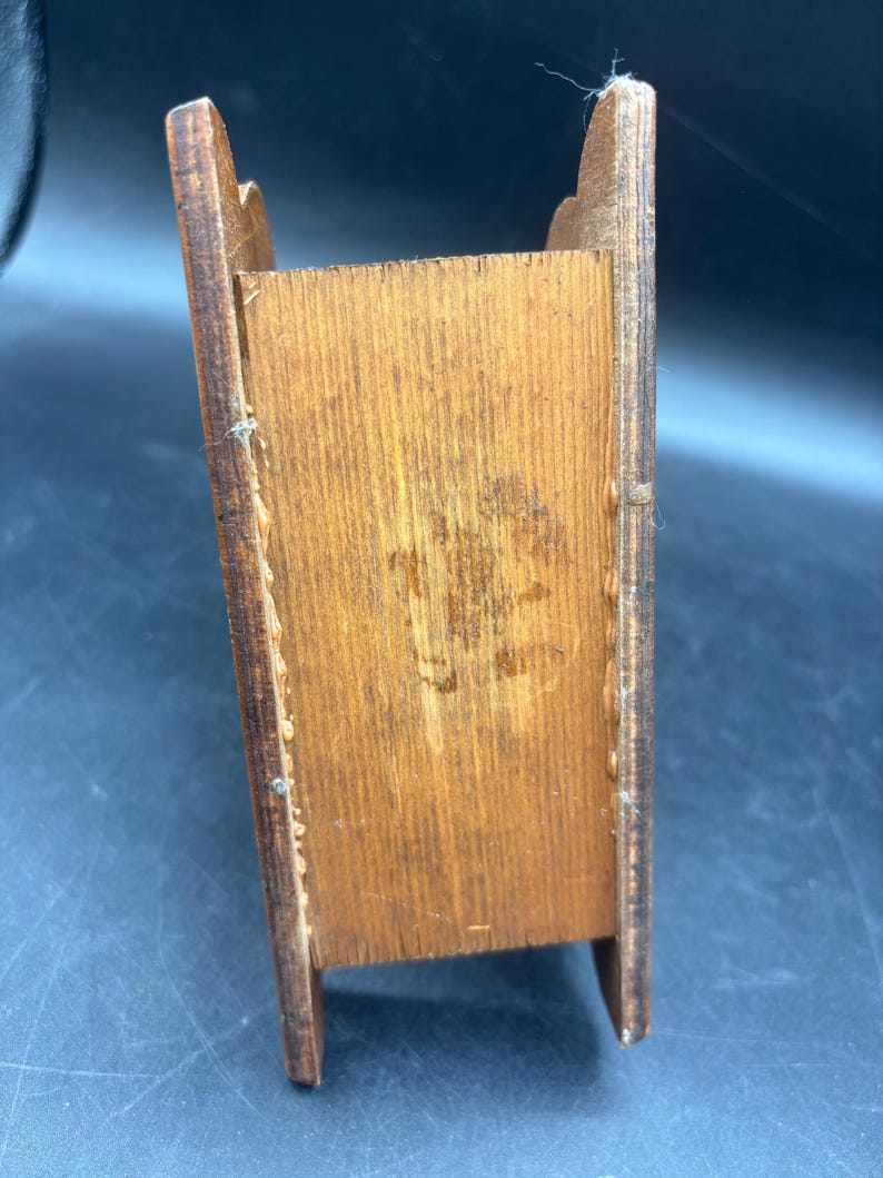 May include: A small, rectangular wooden box with a medium brown finish. The item features two vertical side panels with curved top edges and a central compartment. The wood grain is visible throughout, showing natural variations in color and texture.