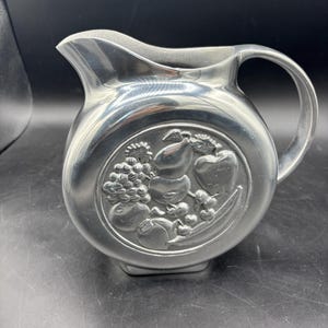 May include: A silver-colored pitcher with a round body and a handle. The pitcher features a raised relief design of various fruits, including grapes, pears, and apples. The pitcher has a spout for pouring and sits on a square base.