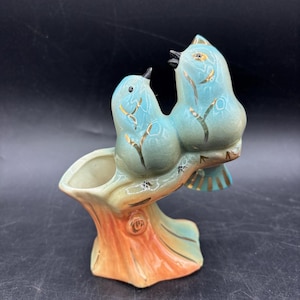 May include: A ceramic planter shaped like a tree stump with two light blue birds perched on top. The birds have gold-colored painted accents on their wings and bodies. The base of the stump is orange and tan, and the interior of the planter is cream.