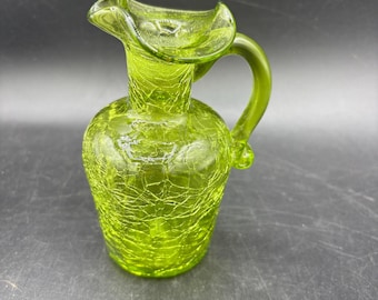 May include: A green glass jug with a crackle finish. The jug has a handle and a ruffled rim. The jug is approximately 15 cm tall.