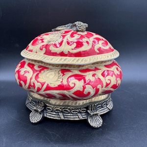 May include: A red and gold ceramic trinket box with a floral design. The box has a lid with a decorative knob and sits on three gold feet.