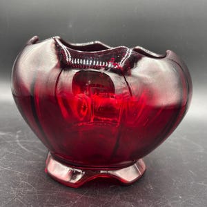 May include: A ruby red glass bowl with a scalloped rim and a pedestal base. The bowl has a rounded shape with vertical ridges and a glossy finish. The glass is transparent, allowing light to pass through and reflect.