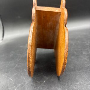 May include: A brown wooden napkin holder featuring two vertical side panels with rounded, scalloped edges connected by a central base piece. The wood grain is visible throughout the structure, showing a natural medium brown tone with darker edges.
