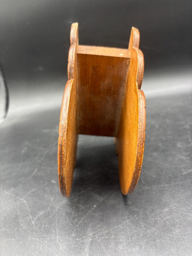 May include: A brown wooden napkin holder featuring two vertical side panels with rounded, scalloped edges connected by a central base piece. The wood grain is visible throughout the structure, showing a natural medium brown tone with darker edges.
