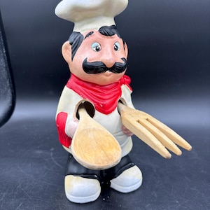 May include: A ceramic chef figurine with a white hat, red neckerchief, and black mustache. The figurine holds a wooden spoon and fork. The chef is wearing a white shirt, black pants, and white shoes.