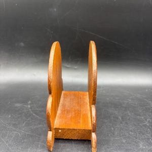 May include: A brown wooden napkin holder featuring two upright, rounded side panels connected by a flat rectangular base. The wood grain is visible throughout the structure, which has a smooth, polished finish and a simple, functional design.