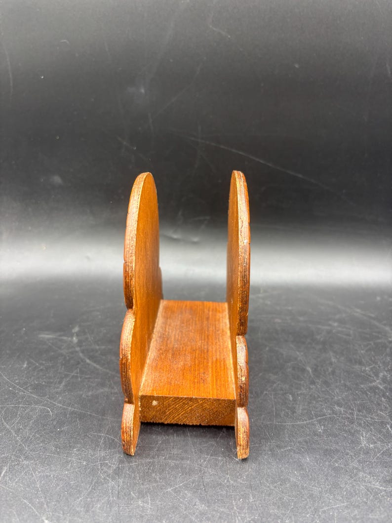May include: A brown wooden napkin holder featuring two upright, rounded side panels connected by a flat rectangular base. The wood grain is visible throughout the structure, which has a smooth, polished finish and a simple, functional design.
