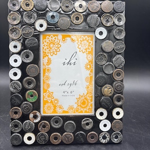 May include: A black photo frame decorated with various metal buttons. The frame holds a 4" x 6" photo and features the text "ihi est 1986 Made in India". The frame has a black border and a floral design.