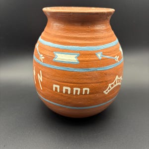 May include: A terracotta-colored clay pot featuring a rounded body and a flared rim. The exterior is decorated with two horizontal light blue painted bands and white hand-painted symbols, including arrows, geometric shapes, and lightning bolts.