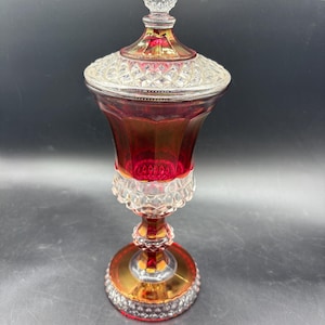 May include: A ruby red and clear glass decorative jar with a lid. The jar has a round base with gold accents and a clear, diamond-patterned lid. The lid is topped with a clear, pointed finial. The jar is likely vintage.