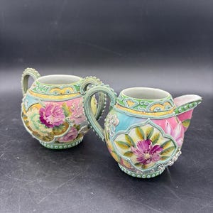 May include: A porcelain sugar bowl and creamer set featuring hand-painted pink and purple floral motifs on a pastel blue and pink background. The pieces are decorated with raised green and gold beaded trim and ornate scrollwork along the rims.