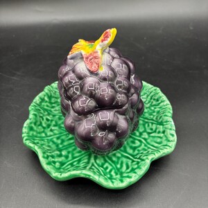 May include: A ceramic grape dish with a green cabbage leaf base. The grapes are purple with a green stem and leaves.