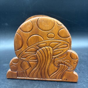 May include: A brown wooden napkin holder featuring a carved relief design of three mushrooms. The largest mushroom is centered with a rounded cap, while two smaller mushrooms are positioned on either side. The wood has a smooth, polished finish.