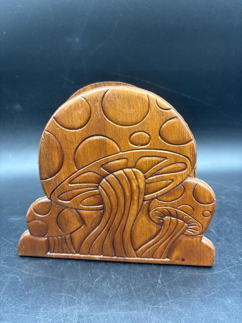 May include: A brown wooden napkin holder featuring a carved relief design of three mushrooms. The largest mushroom is centered with a rounded cap, while two smaller mushrooms are positioned on either side. The wood has a smooth, polished finish.