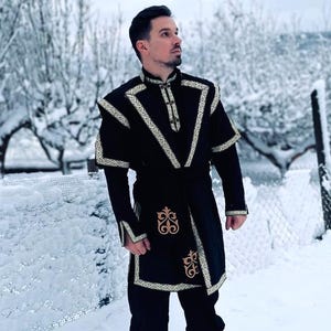 Könnte beinhalten: Schwarze Tunika mit goldfarbenen Verzierungen und dekorativen Stickereien. Die Tunika wird mit einer schwarzen Hose und Stiefeln getragen. Das Outfit wird in einer verschneiten Umgebung im Freien getragen. Die Tunika hat einen V-Ausschnitt und ist an der Taille gegürtet.