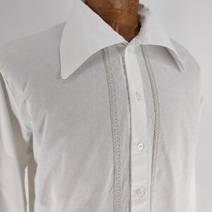 May include: White button-down shirt with a large collar and long sleeves. The shirt features decorative lace trim down the front and is buttoned up. The shirt is displayed on a mannequin.