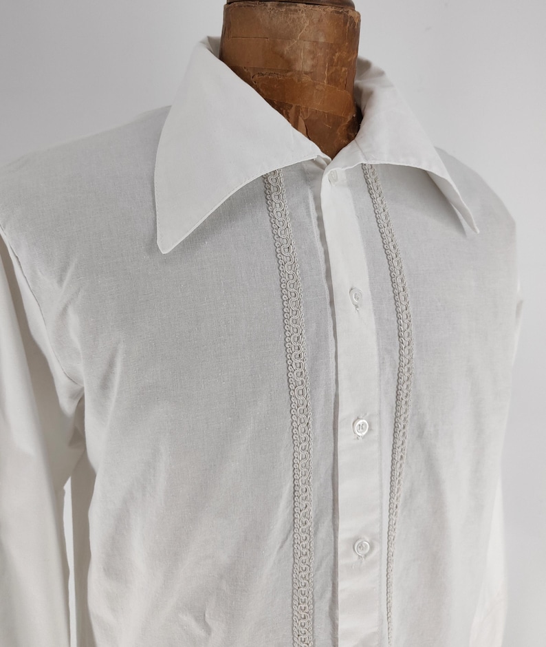 May include: White button-down shirt with a large collar and long sleeves. The shirt features decorative lace trim down the front and is buttoned up. The shirt is displayed on a mannequin.