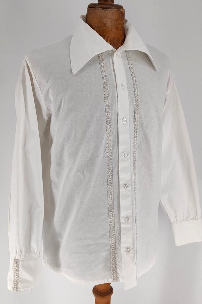 May include: A white long-sleeve shirt with a wide collar and button-up front. The shirt features decorative vertical lace trim on either side of the button placket and on the cuffs. The shirt is displayed on a wooden mannequin.