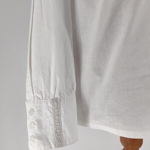 May include: A white long-sleeved shirt with a buttoned cuff. The cuff features three small buttons and a decorative trim. The shirt is made of a lightweight fabric, and the sleeve is slightly wrinkled.
