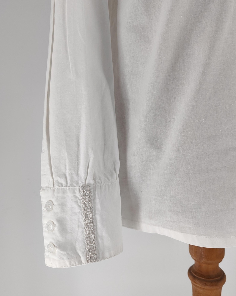 May include: A white long-sleeved shirt with a buttoned cuff. The cuff features three small buttons and a decorative trim. The shirt is made of a lightweight fabric, and the sleeve is slightly wrinkled.
