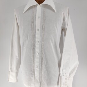 May include: A white long-sleeve shirt with a wide collar and button-up front. The shirt features decorative vertical lace trim on the front and cuffs. The cuffs have three small buttons. The shirt is displayed on a wooden mannequin.