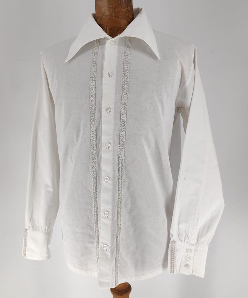 May include: A white long-sleeve shirt with a wide collar and button-up front. The shirt features decorative vertical lace trim on the front and cuffs. The cuffs have three small buttons. The shirt is displayed on a wooden mannequin.