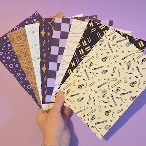 May include: A hand displays a selection of patterned papers against a purple backdrop. The papers feature diverse designs, including dots, stars, and geometric shapes in purple, gold, and black. Patterns include a checkerboard and a design with pens and whistles.