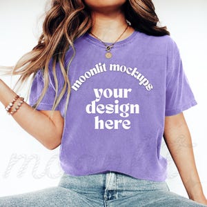 May include: A lavender t-shirt with the text "moonlit mockups your design here" in white. The model is wearing light wash denim jeans and white sneakers. A gold necklace and bracelet are also visible.