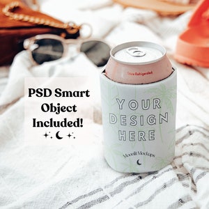 Puede incluir: Enfriador de latas blanco con un diseño tropical y el texto "YOUR DESIGN HERE". El enfriador está sobre una toalla blanca y gris a rayas con un par de sandalias rosas.