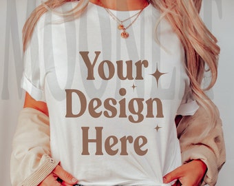 May include: A white t-shirt with the text "Your Design Here" in brown lettering. The shirt is being worn by a person with long blonde hair and a light brown jacket. The person is wearing blue jeans with a distressed knee.