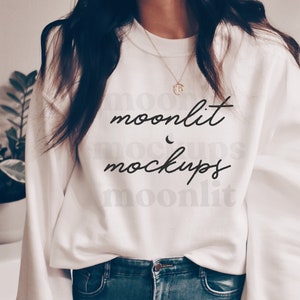 May include: A white sweatshirt with the text "moonlit mockups" in black script. The sweatshirt is worn by a person wearing blue jeans.