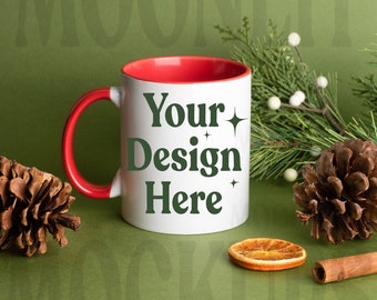 May include: A white ceramic mug with a red interior and handle, featuring the text "Your Design Here" in green. The mug is surrounded by pine cones, greenery, a dried orange slice, and a cinnamon stick, set against a green background.